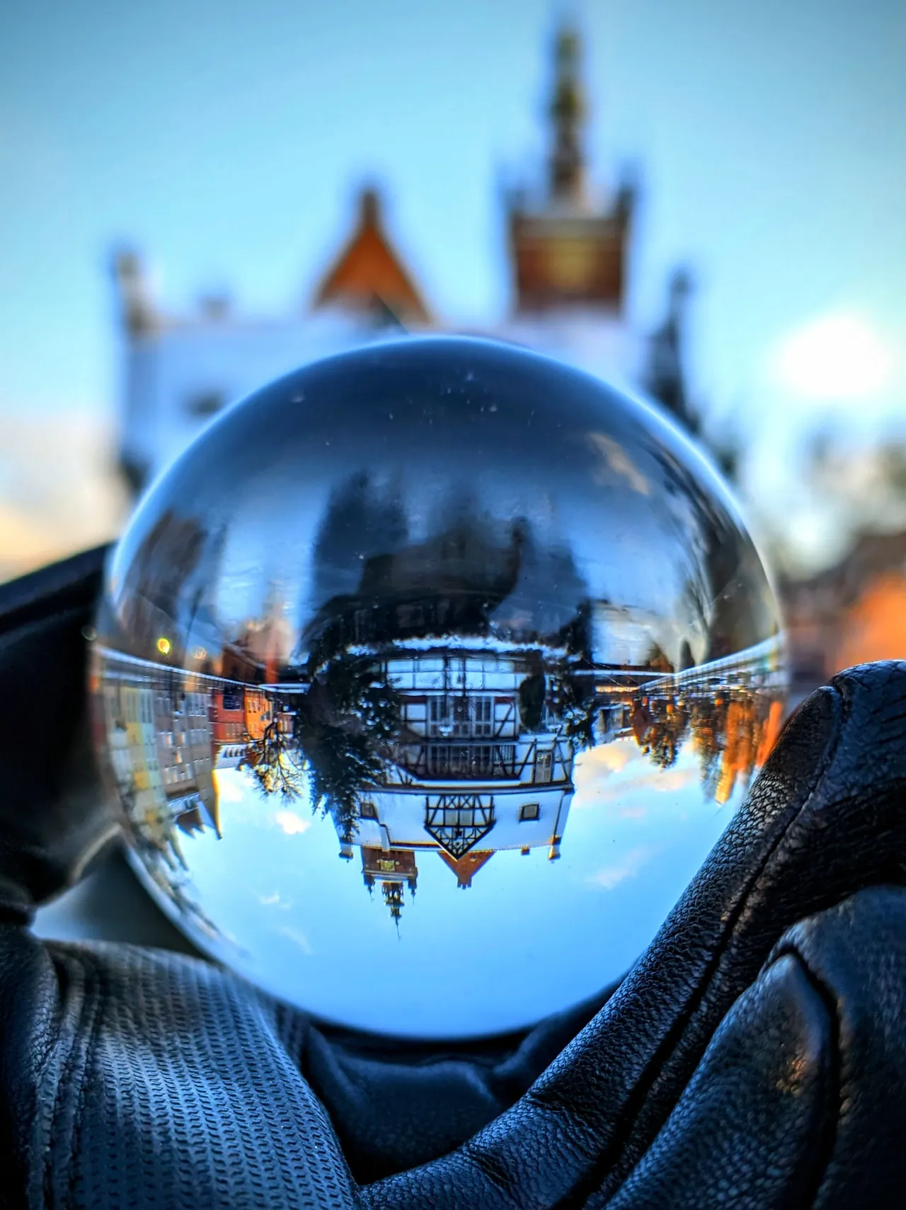 A glass ball held in a gloved hand, showing the half-timbered Millers' Guild Hall in Gdańsk upside down, with the building itself blurred behind it against a blue sky.