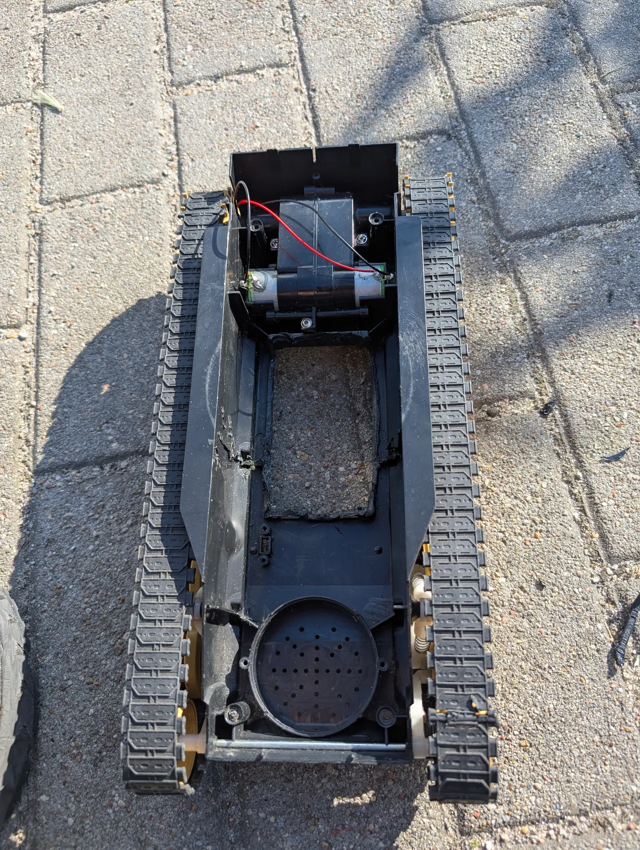 The open hull of a toy tank on paving stones in the sun: an empty plastic shell on two tracks, with only a motor and gearbox and a red wire left at one end.