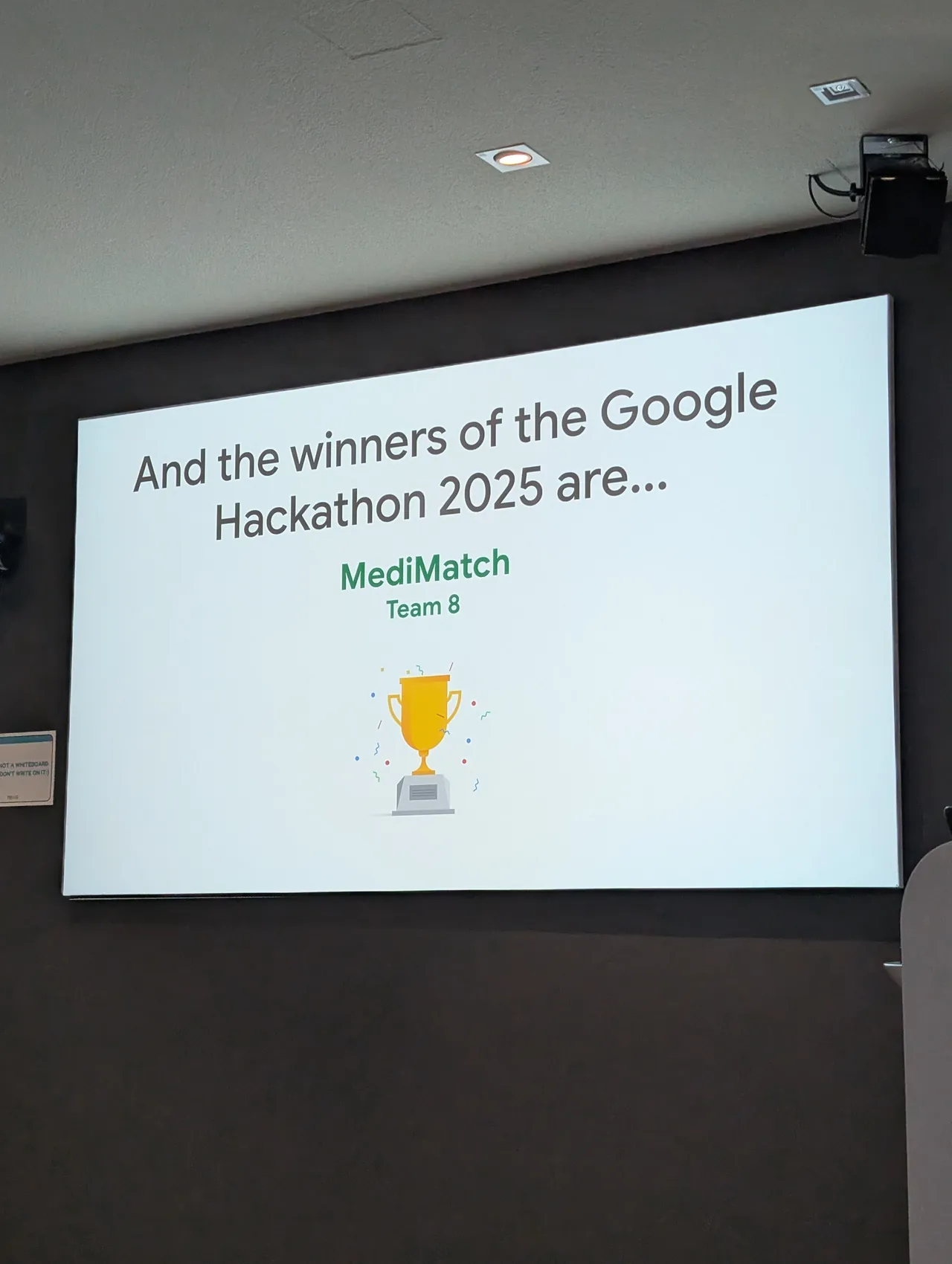 A projector screen reading 'And the winners of the Google Hackathon 2025 are... MediMatch, Team 8', above a gold trophy.