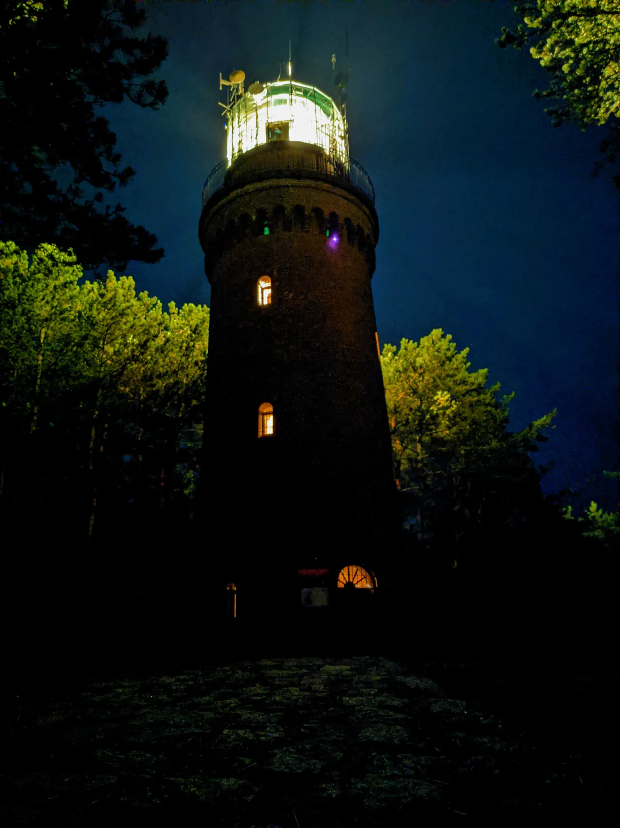 A round brick lighthouse at night, its lamp room lit, with lit windows lower down and trees on both sides against a dark blue sky.