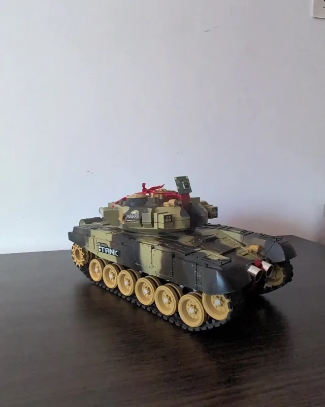 A camouflage toy tank on a dark table against a white wall, a small camera board tied to its turret with red ribbon and an ultrasonic sensor on its front.