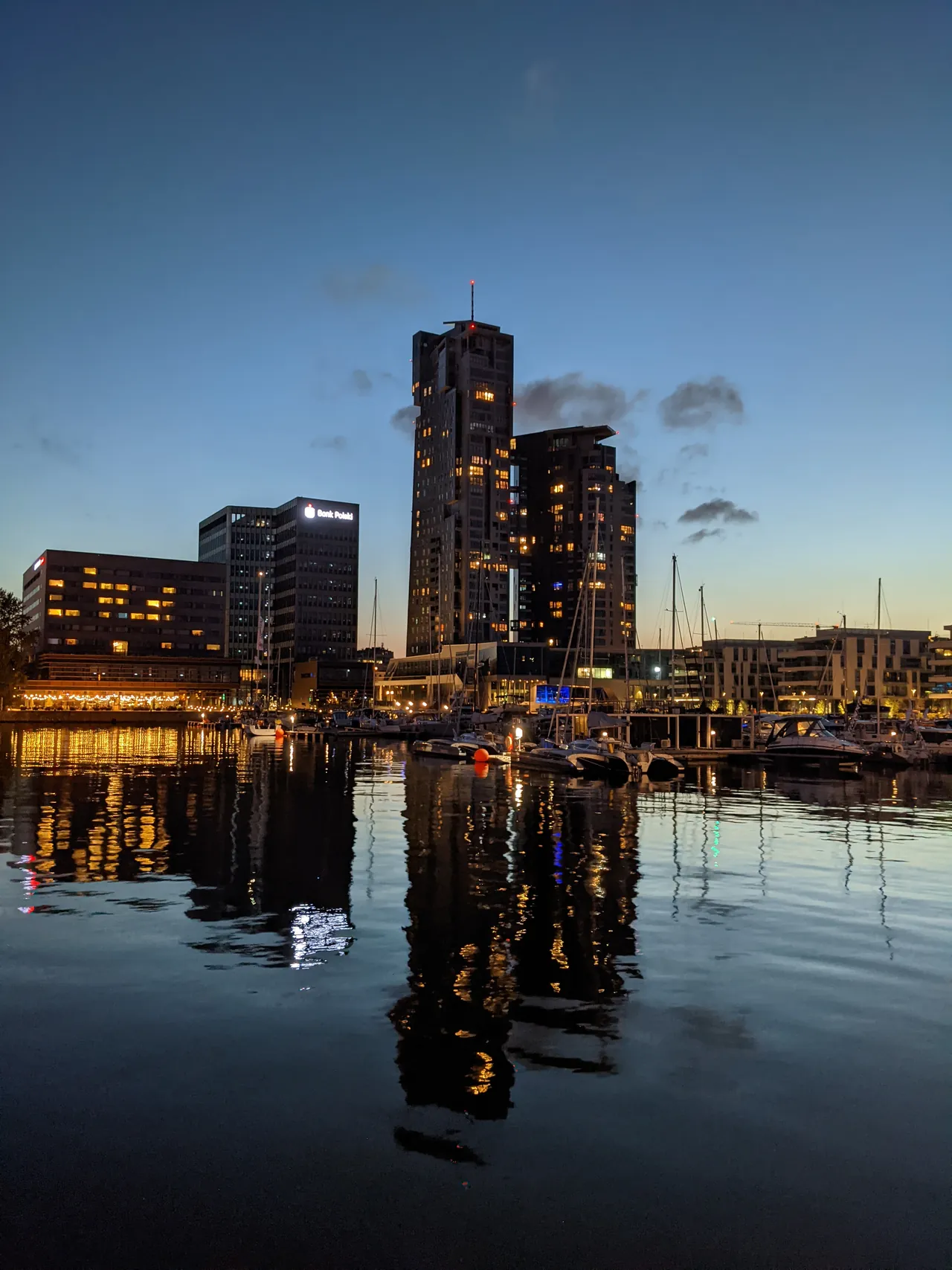 Dark towers and an office block at dusk above a marina, their lit windows and masts reflected in calm water.