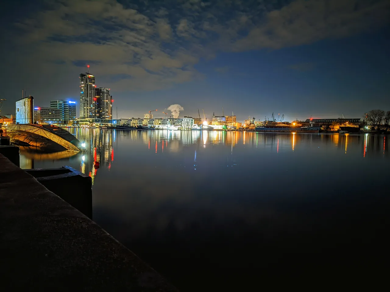 A still harbour at night seen from a stone quay, a submarine's hull at the left; tall towers and lit buildings on the far shore, port cranes, and their lights reflected in the water.