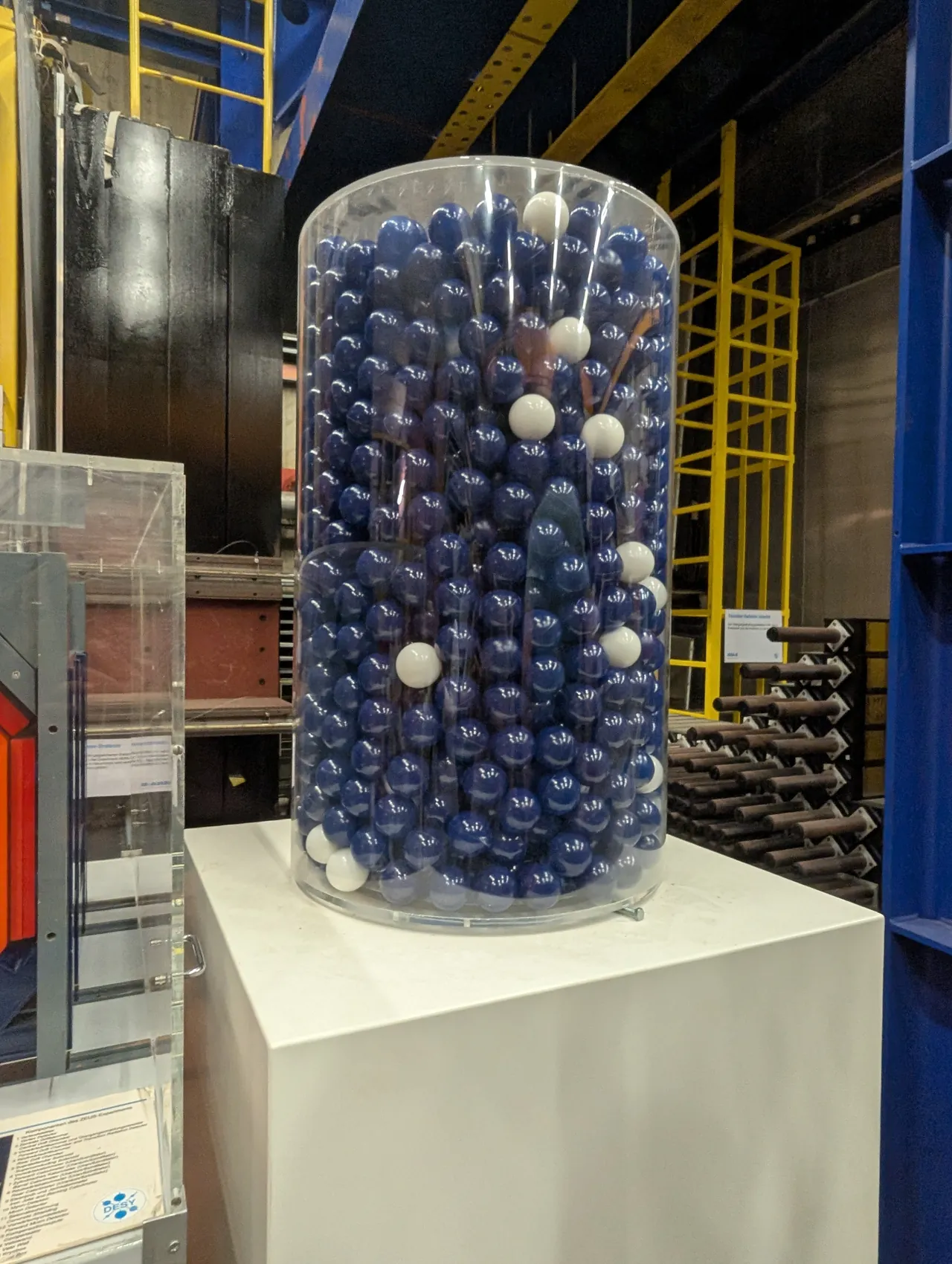 A tall clear cylinder filled with dark blue balls and a few white ones, on a white plinth in a hall of yellow steel ladders at DESY.