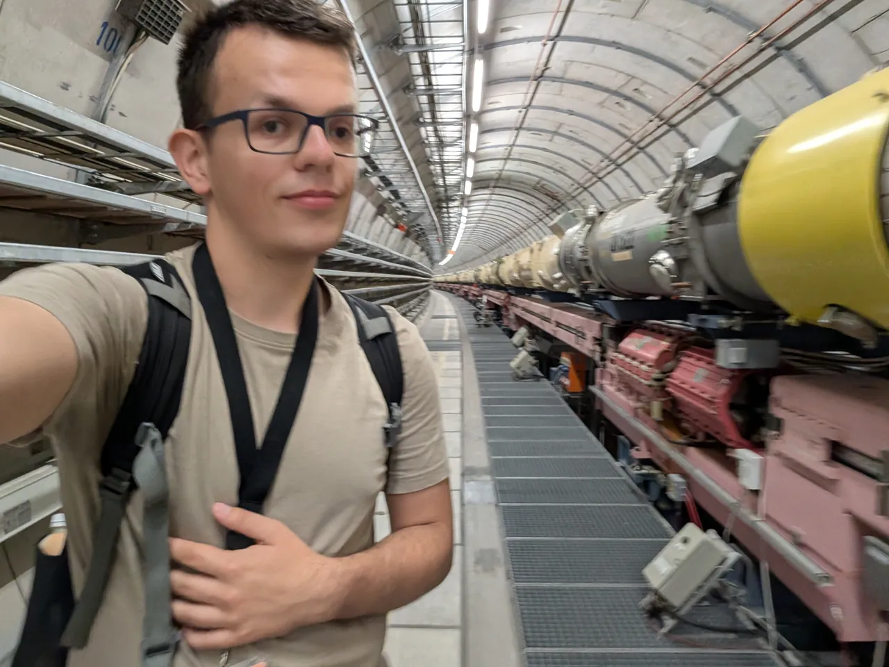 Wojciech taking a selfie in an accelerator tunnel at DESY, the yellow and red magnets of the beam line running into the distance along the curved concrete wall.