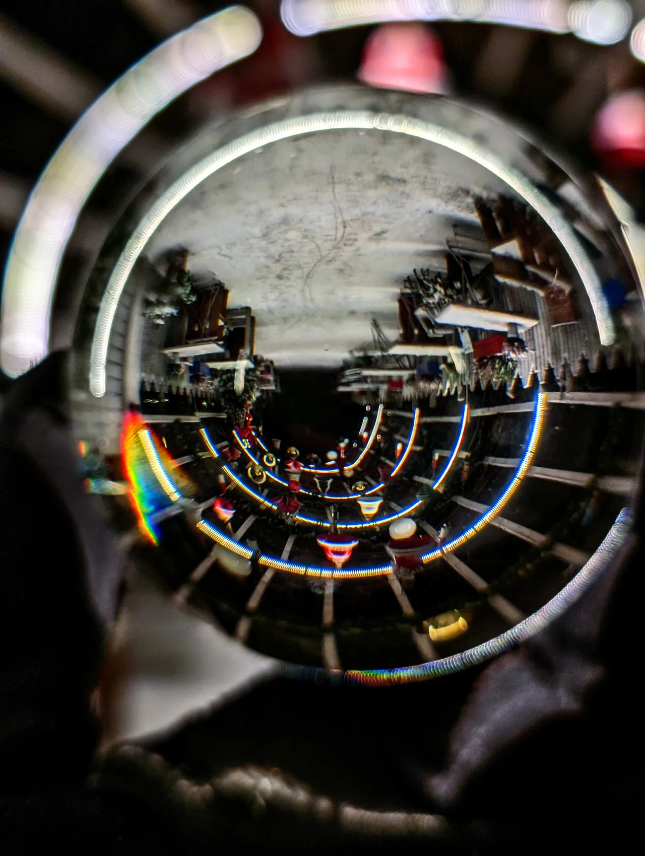 A glass ball held close to the camera; inside it a space with shelves and string lights is turned upside down and bent into glowing rings, with a rainbow at the edge of the glass.